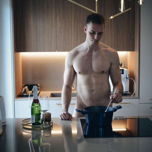 The photo shows Erik standing, preparing lunch. He is completely naked. His crotch is covered by a black pot. Erik is a very handsome blond with large breasts and defined abs. He is holding a ladle in his left hand and stirring the cooked lunch. In his right hand is a bottle of champagne with 2 glasses. In the back is a coffee machine and a lit kitchen unit. Daylight comes from the left side, which nicely highlights Erik's muscles and completes the sensuality of the photo.