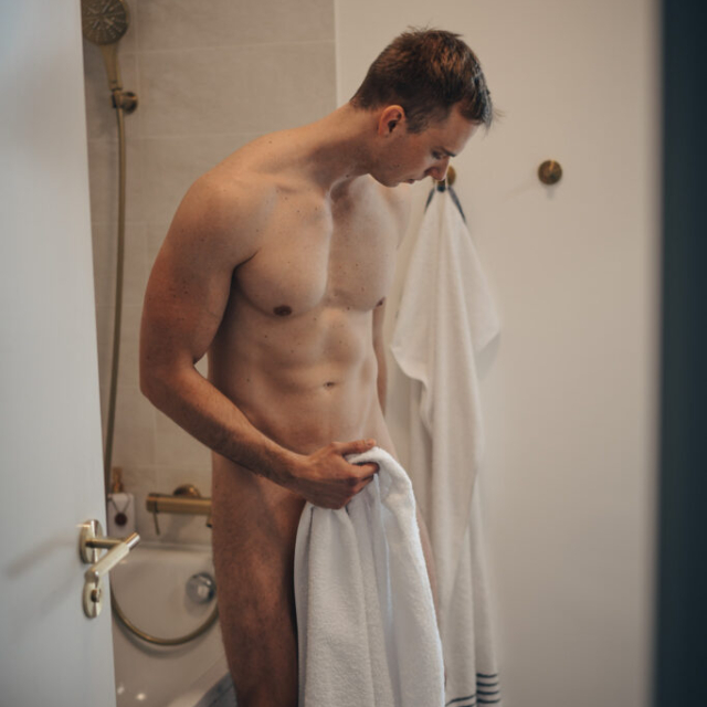 The photo is taken from the hallway and shows a naked Erik in the bathroom, stepping out of the bathtub after a shower. Erik is drying himself with a white towel and looking down. The towel covers his crotch. In the background is a white ceramic bathtub with gold accessories. Daylight illuminates the model from the right, highlighting his perfect masculinity.