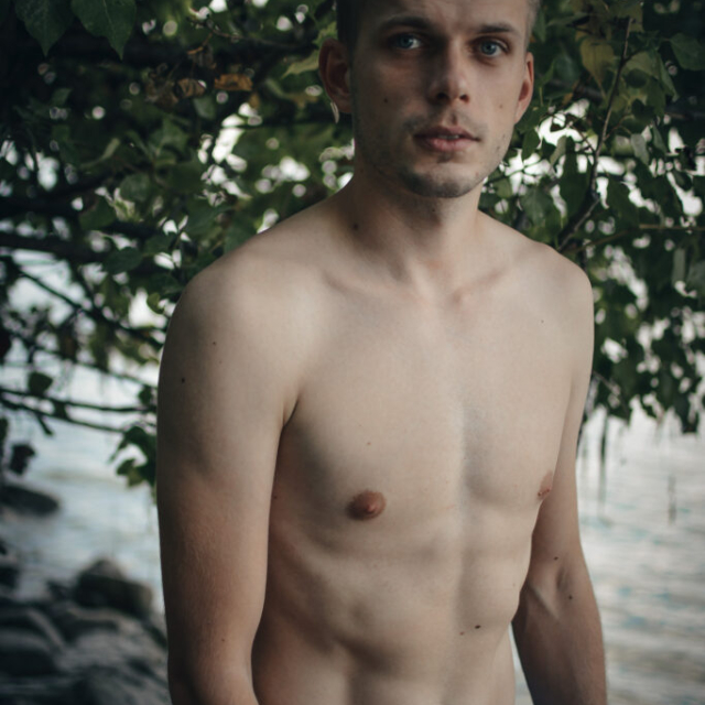 In the photo, Michal R is on the bank of the Danube River. He has short blond hair and a smooth body. He is standing on the stones in blue Kelvin Clein boxer shorts. In the background, thickets of bushes and a flowing river can be seen. The cloudy weather completes the atmosphere of this portrait photo.