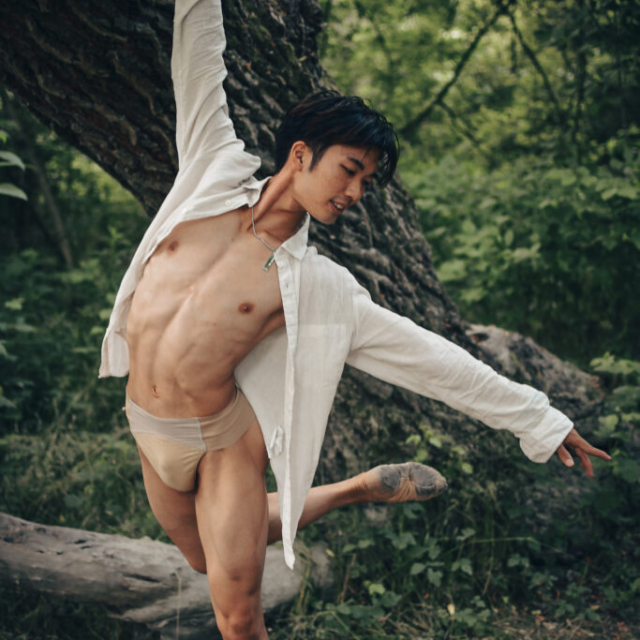 Dans is photographed in a ballet pose in a dense deciduous forest on the banks of the Danube. He is a young Asian man, with black hair. He is wearing an unbuttoned white shirt, dance underwear and ballet shoes. The perfect dance pose in harmony with nature enhances the atmosphere of the photo. In the background is the thick trunk of a deciduous tree and a thicket of bushes, branches and leaves.