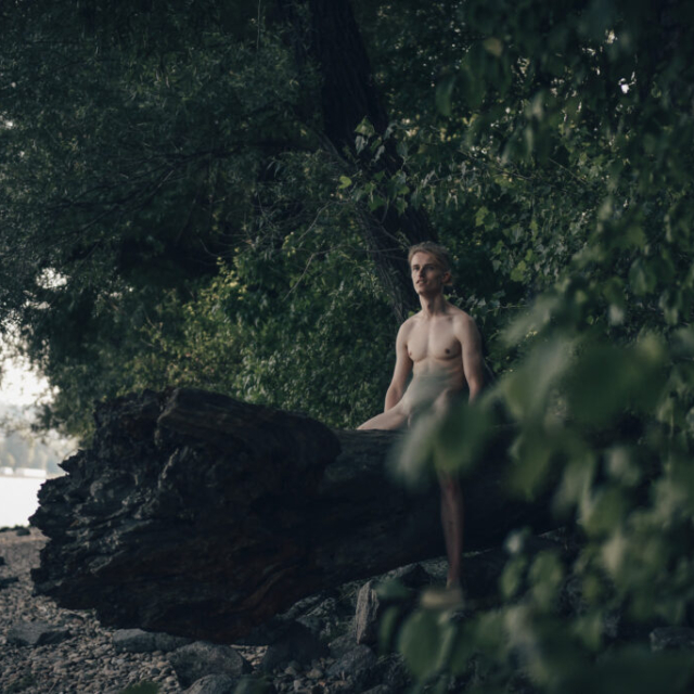 A sensual portrait of an athletic male model, Vasilij, who sits timidly on a large stump of a fallen tree with his legs spread wide. The photo is set in the Bratislava riverside setting. Vasilij is a blond, with nicely developed breasts. He has long hair tied in a ponytail. He is looking towards the Danube River. Below him is a rocky beach and on the left side the river flow. His cock is subtly visible through the tree foliage, which completes the sensuality of this photo.