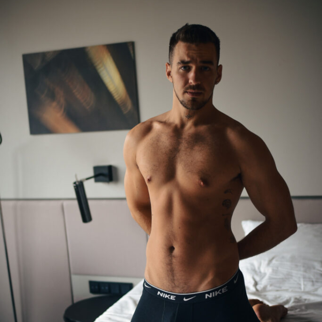A sensual morning portrait of a muscular, shirtless man named David B. kneeling on a bed in a Berlin hotel room. He wears black Nike boxers, holding his hands behind his back and looking at the camera.