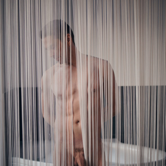 An artistic, sensual portrait of a naked man named Lucas, viewed through delicate, vertical curtains. He looks down thoughtfully, his legs spread, his large penis protruding through the curtain, in a Berlin hotel room, with a white bathtub partially visible in the background.