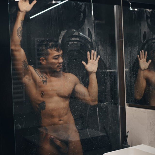A moody, sensual portrait of a muscular, tattooed man named Andrew standing in a dark shower at Eurovea Tower in Bratislava. He is looking away while pressing his hands against the wet glass enclosure.