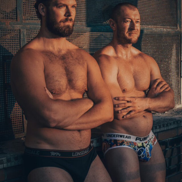 A sensual NSFW portrait of two shirtless men, Juraj and Martin, wearing briefs. They stand with crossed arms at an abandoned stadium in Brno, illuminated by warm golden hour sunlight.