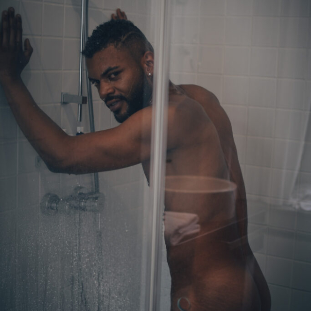 A moody, sensual NSFW portrait of a shirtless, muscular man named Kevin standing under a running shower. He is leaning forward, pressing both hands flat against the white tiled shower wall. He has dark curly hair and a beard, and is looking back over his right shoulder directly at the camera with an intense expression. The image is shot through a partially visible glass shower door, with water droplets adding texture to the dim, cinematic lighting.