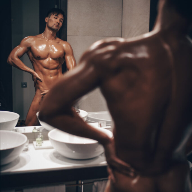 A detailed sensual NSFW portrait of a shirtless, oiled, muscular Asian man named Will, with a mustache and goatee, standing in a dimly lit bathroom. He is seen directly from behind. He is posing in front of a large, dark-framed mirror which provides a full frontal reflection of his body and face. His front is glistening with oil, highlighting his defined abs and chest. Moody ambient light illuminates his defined physique.