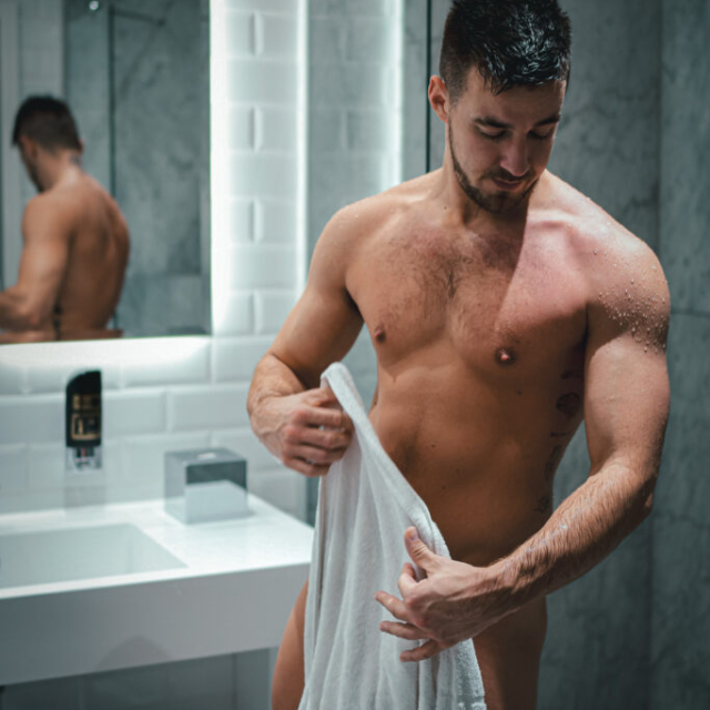 A sensual NSFW portrait of a muscular, naked man named David D. standing in a modern bathroom. He has short wet dark hair, a beard, and chest hair. Water droplets are visibly glistening on his left shoulder and arm. He is looking down with a thoughtful expression while holding a white towel with his right hand. Behind him, a mirror framed by bright vertical LED lights reflects his bare back. The bathroom features white subway tiles and elegant grey marble-patterned walls. A blurred black bottle sits in the lower left foreground.