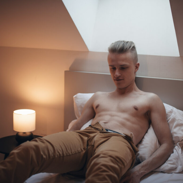 A sensual portrait of a shirtless man named Vasilij with a blonde undercut hairstyle. He is lying back on a bed against white pillows, looking downwards with a relaxed expression. He is wearing khaki trousers that are casually unbuttoned at the waist. The bedroom features a sloped ceiling with a skylight directly above him. To his right (viewer's left), a small cylindrical lamp glows warmly on a round black nightstand, casting a moody, soft light across his torso and the room.