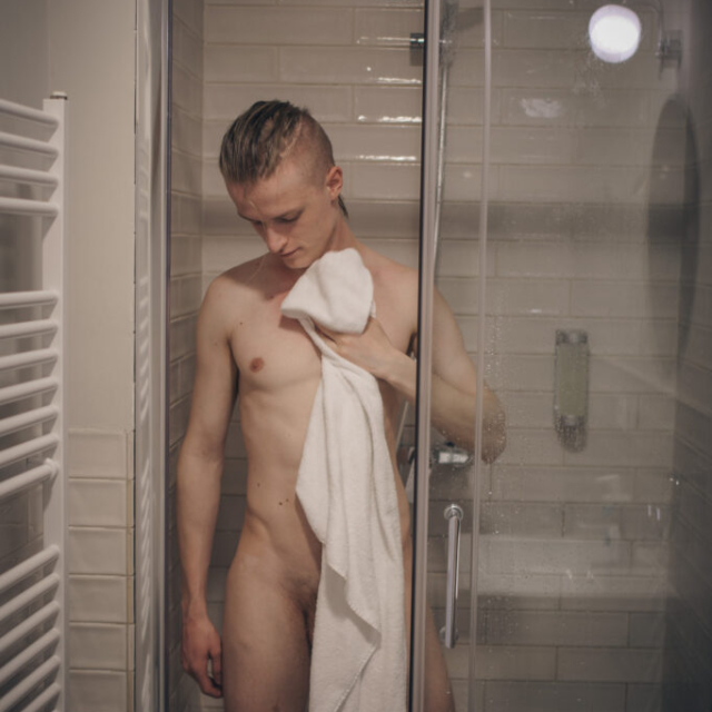 Sensual portrait of a shirtless young man with blond hair tied in a ponytail in a modern bathroom. He is standing in a glass shower stall and drying himself with a white towel. He is looking at his wet naked body.