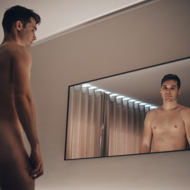 Artistic portrait of a male model standing in front of a large mirror in a hotel room. Exclusive uncensored premium gallery teaser.