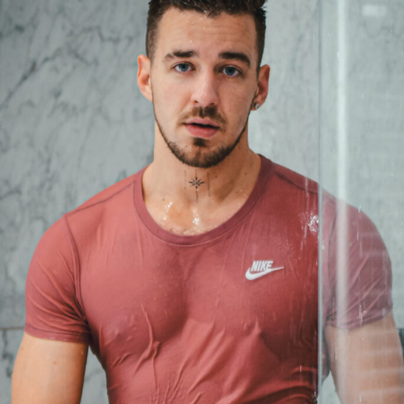 A man stands under a running shower wearing a white t-shirt that clings to his skin. Artistic Water VIP Series.