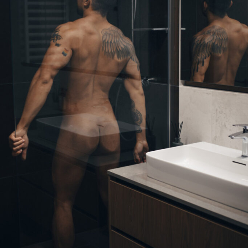 A muscular man with tattoos stands with his back pressed against the glass of a shower. Noir Collection.