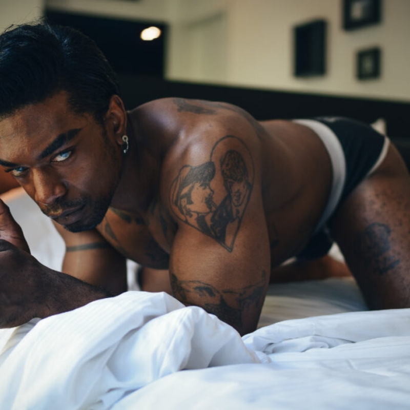 A Black man kneels on a bed. He leans forward and looks at the camera with very bright blue eyes. VIP access preview.