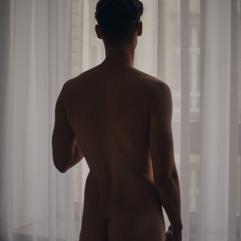 A athletic man stands before a sheer curtain, gazing through the fabric and window at the world outside. Insider premium look.