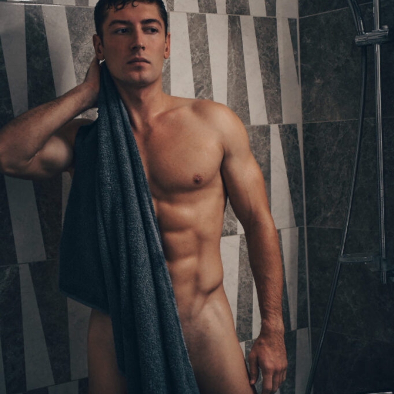 Muscular male model with a towel over his shoulder standing in a modern shower. High-quality premium male aesthetics.