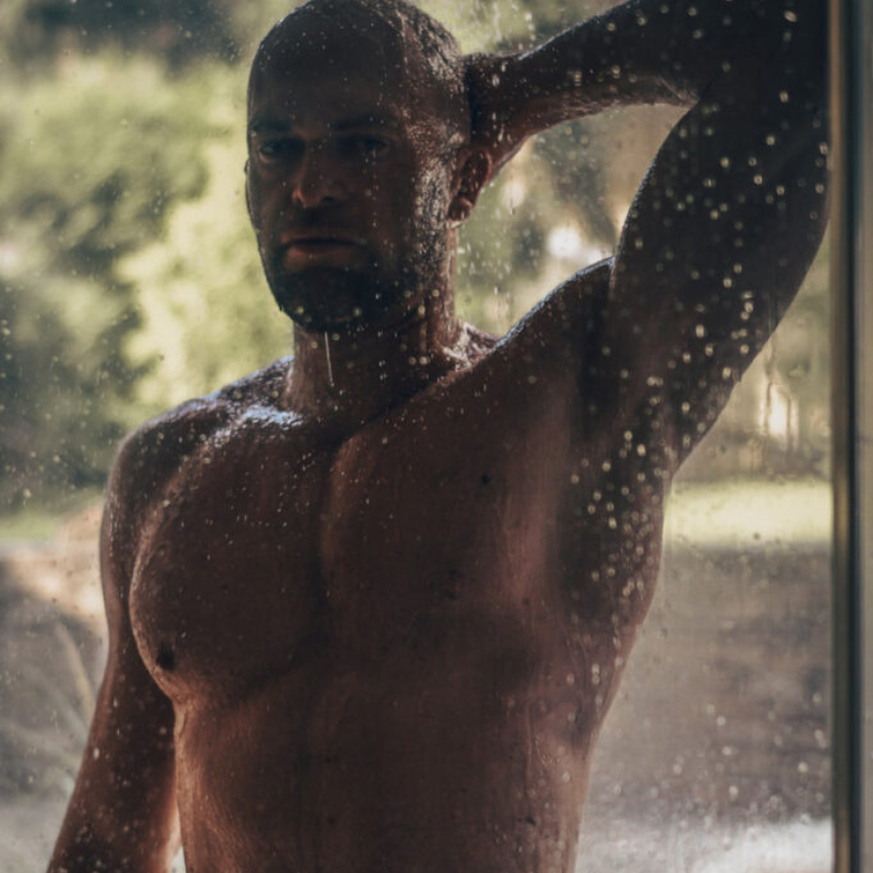 Muscular and masculine male model taking a shower behind wet glass. Exclusive gay erotic photography and cinematic lighting.