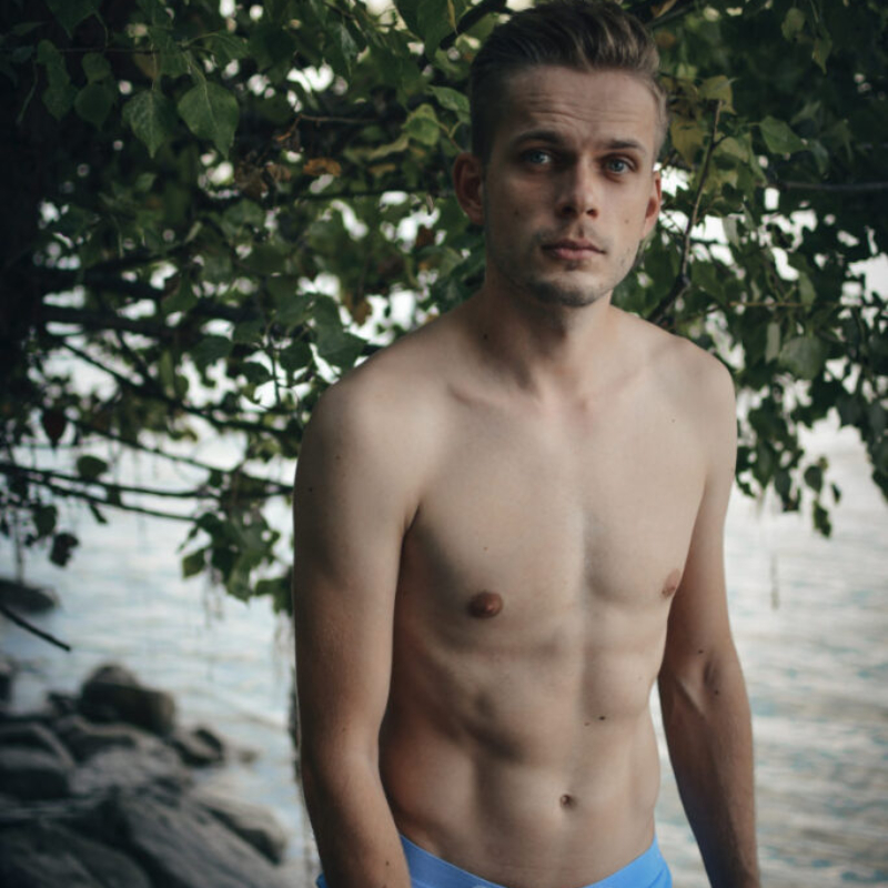 An athletic young man stands by the Danube river. He looks directly at the camera with a shy expression. VIP Only Glance.