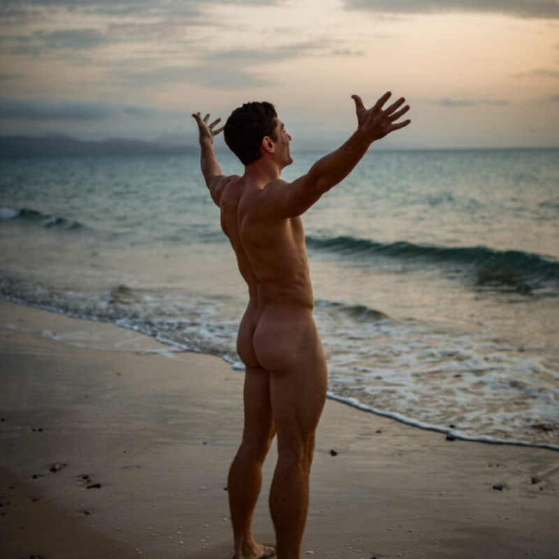 Artistic outdoor portrait of a fit male model standing on a sandy beach with arms raised at sunrise. Sensual and exclusive male aesthetics.