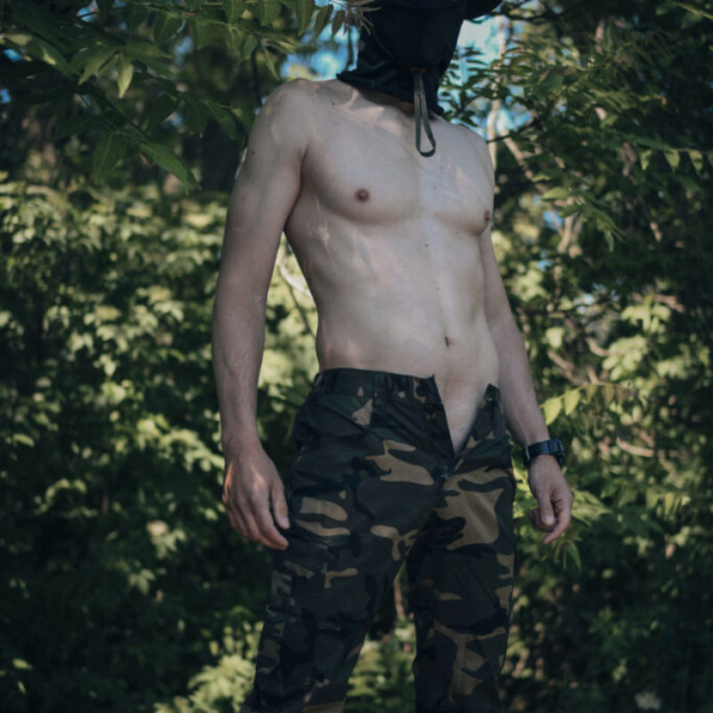 A powerful outdoor photo of a fit, muscular man posing on a concrete military bunker near the Danube river in Bratislava. He is wearing camouflage trousers and a matching army hat. He shows his highly defined abs and athletic body.