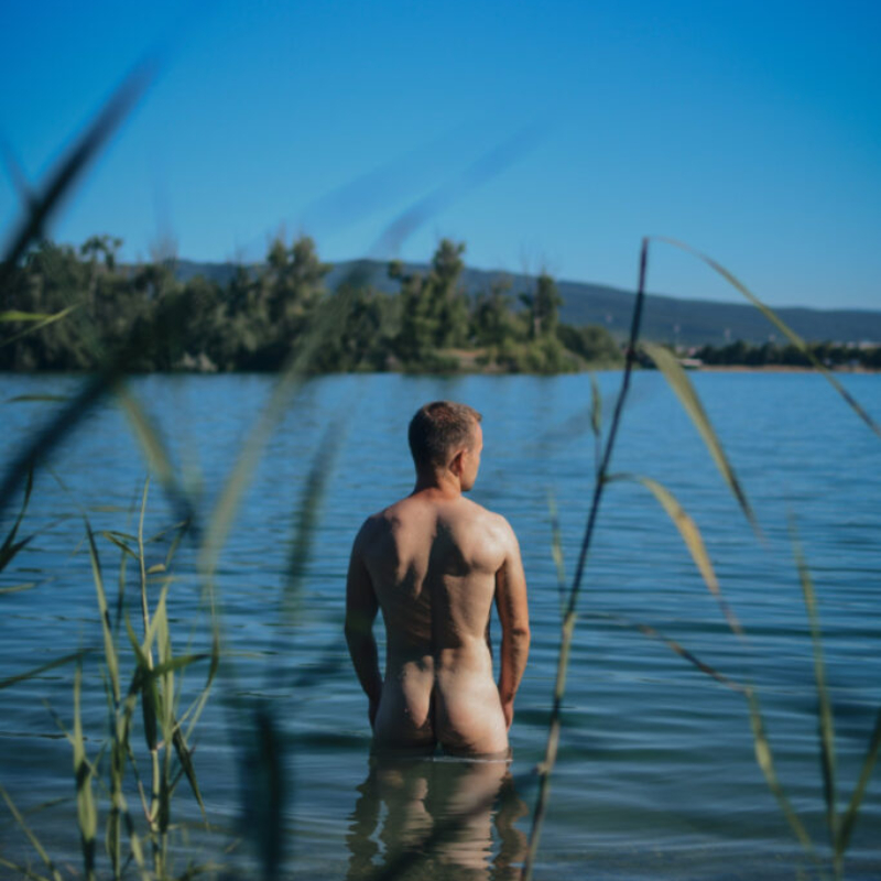A young man stands in Zlate Piesky lake, seen through tall grass during a bright, sunny afternoon.