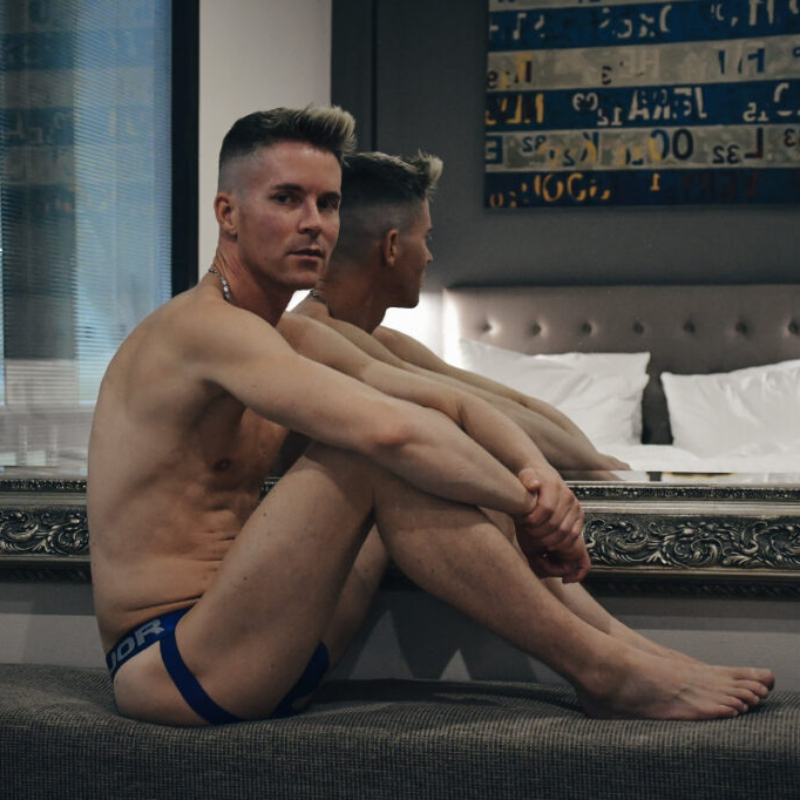 Fit male model wearing athletic underwear sitting gracefully in a hotel bedroom. Uncensored premium male photography preview.