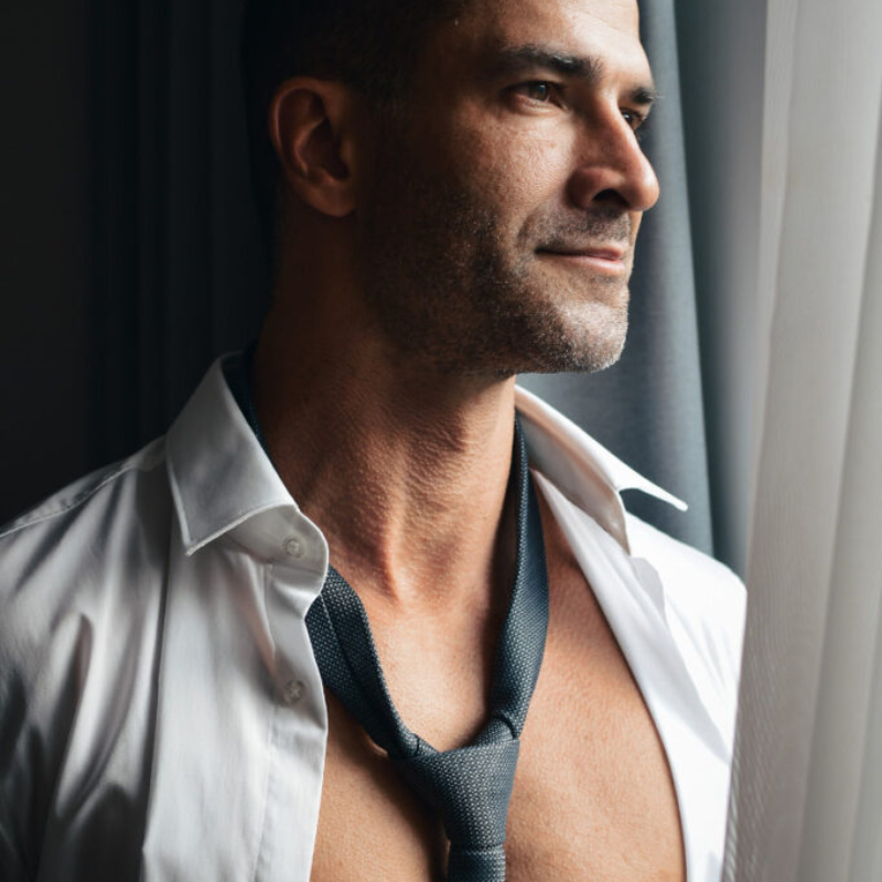 Attractive mature male model with an open white shirt and loose tie looking out the window. Premium lifestyle male aesthetics.
