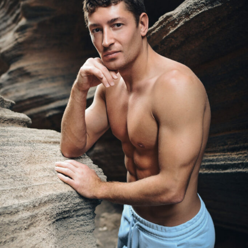 Attractive athletic male model in light blue shorts posing confidently among natural rock formations. High-quality outdoor male aesthetics.