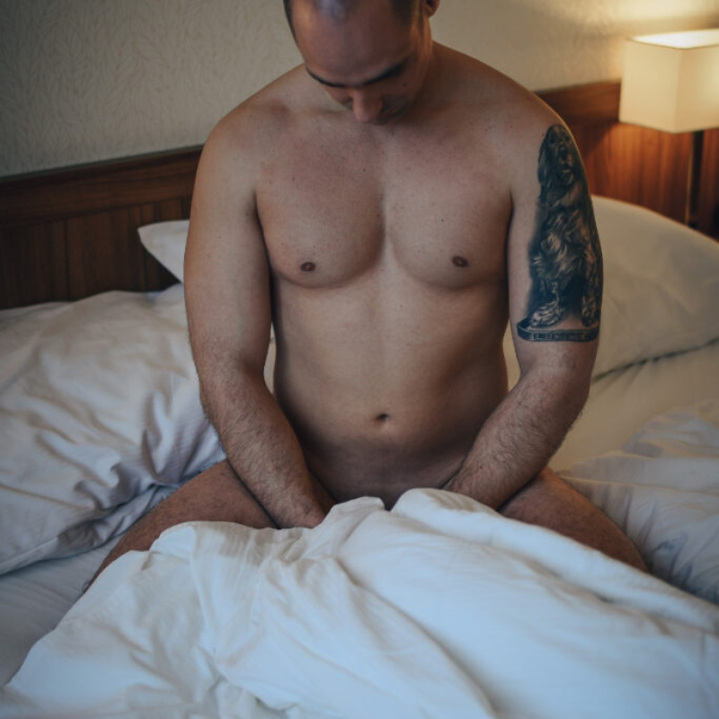 Sitting on his heels, the young man lowers his head to observe his chest, creating a moody, introspective bedroom scene. Insight to Premium Access.