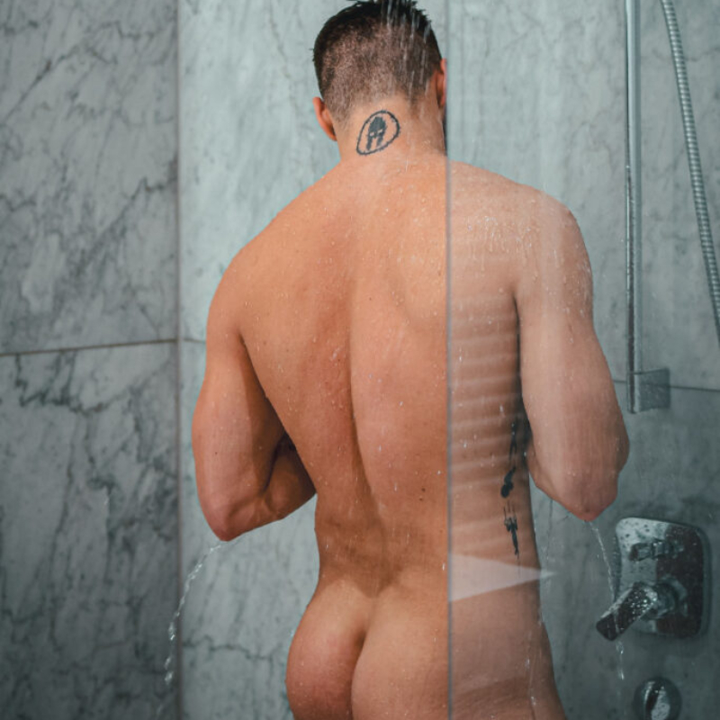 A man stands with his back to the camera, washing himself under a steady stream of water in the shower. VIP All-Access view.