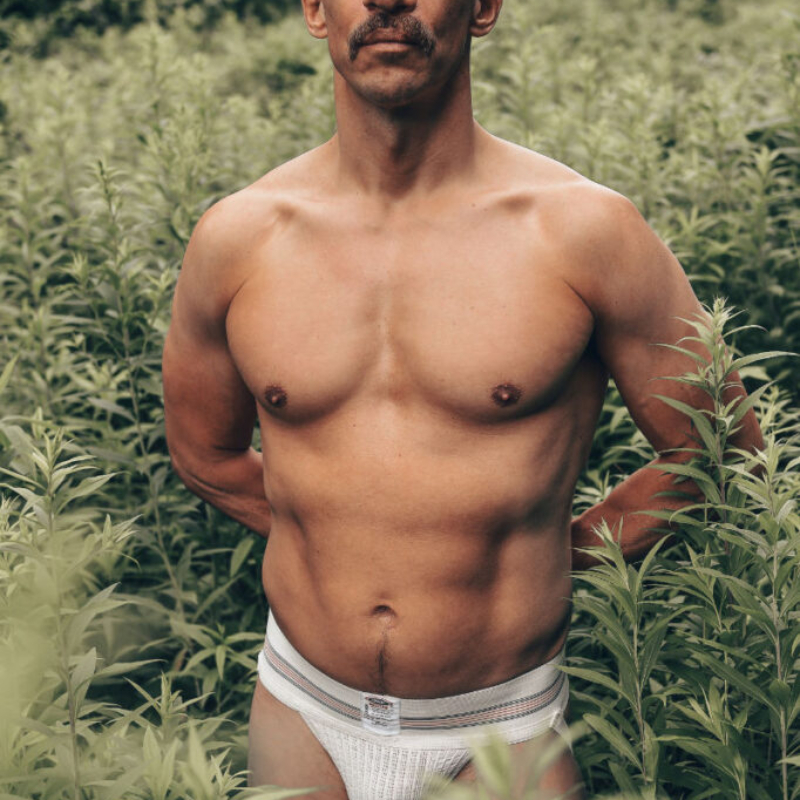 Athletic male model wearing a white jockstrap standing in tall forest grass. Bold and exclusive outdoor male aesthetics.