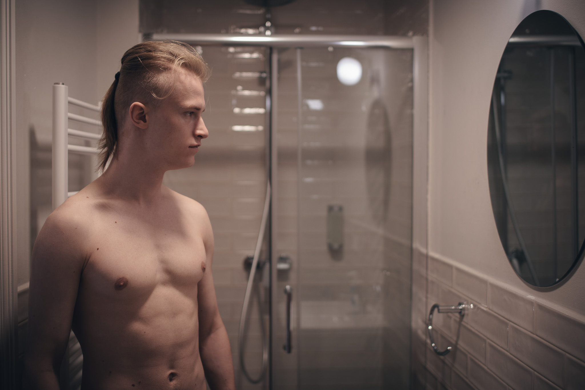 A sensual portrait of a shirtless young man with blonde hair tied in a ponytail, standing in a modern bathroom. The background features a glass shower cabin and a white radiator, with a round mirror visible on the right.