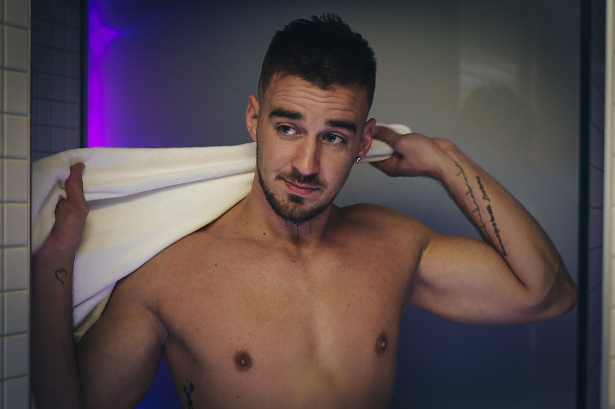 A man with a towel wrapped around him looks toward the mirror with a playful, flirty expression. Members Only Gallery Preview.