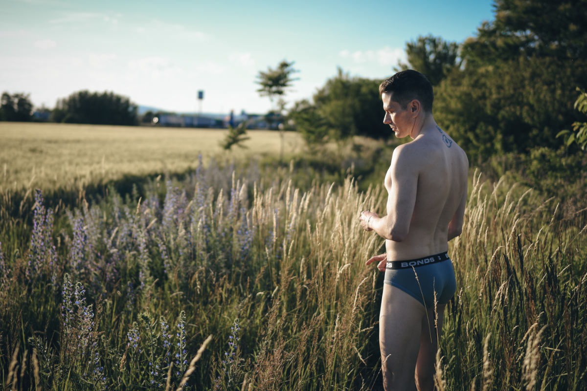 Man standing in a lush green meadow, watching a vibrant sunset during the golden hour. Warm sunlight illuminates the wild flowers and high grass around him as he looks into the distance.
