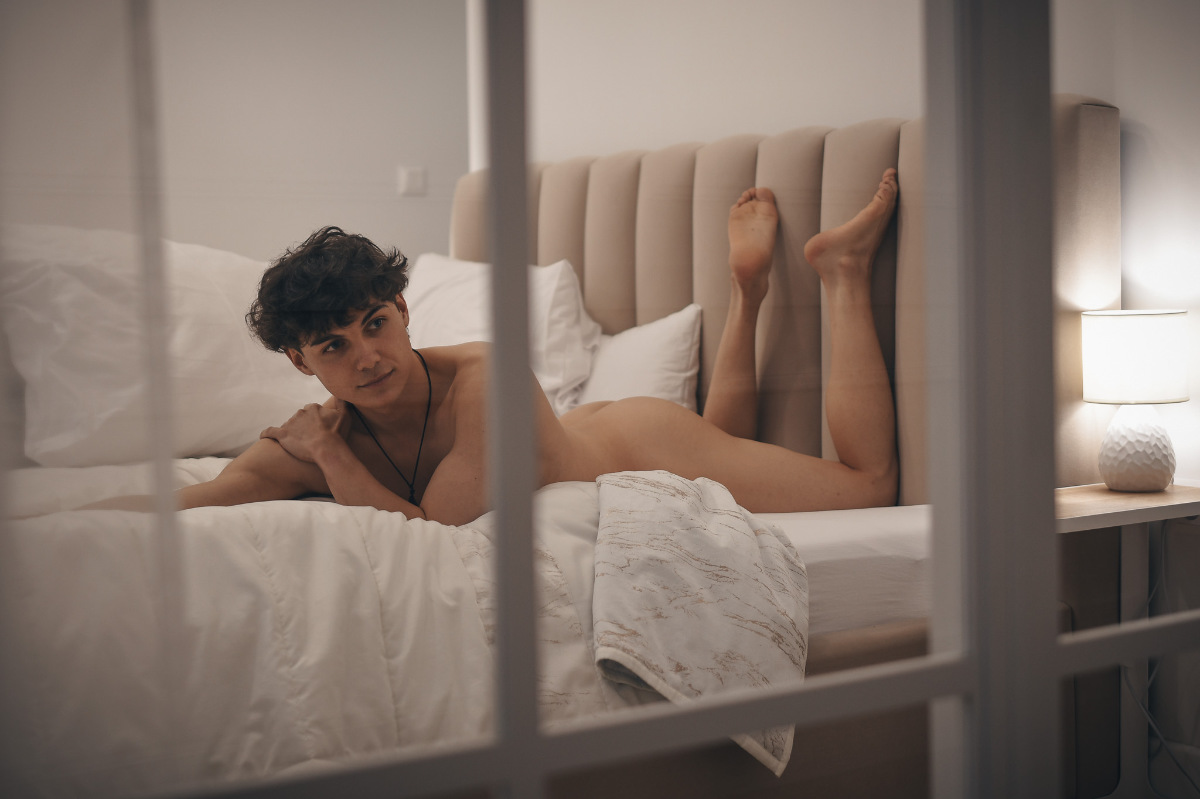 Twink on the bed, captured through the glass, which allows you to see directly into the bedroom. He is on his stomach and resting. He has nice curly hair. The atmosphere is completed by the interior lights.