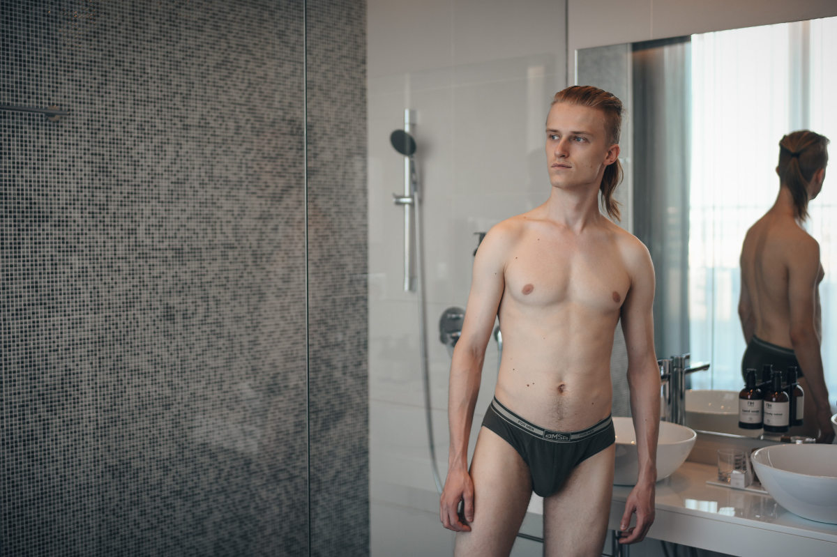 A blonde male model with a ponytail stands in a modern bathroom wearing dark grey briefs. He is looking away from the camera, next to a glass shower wall and a white sink. His reflection is visible in the mirror.