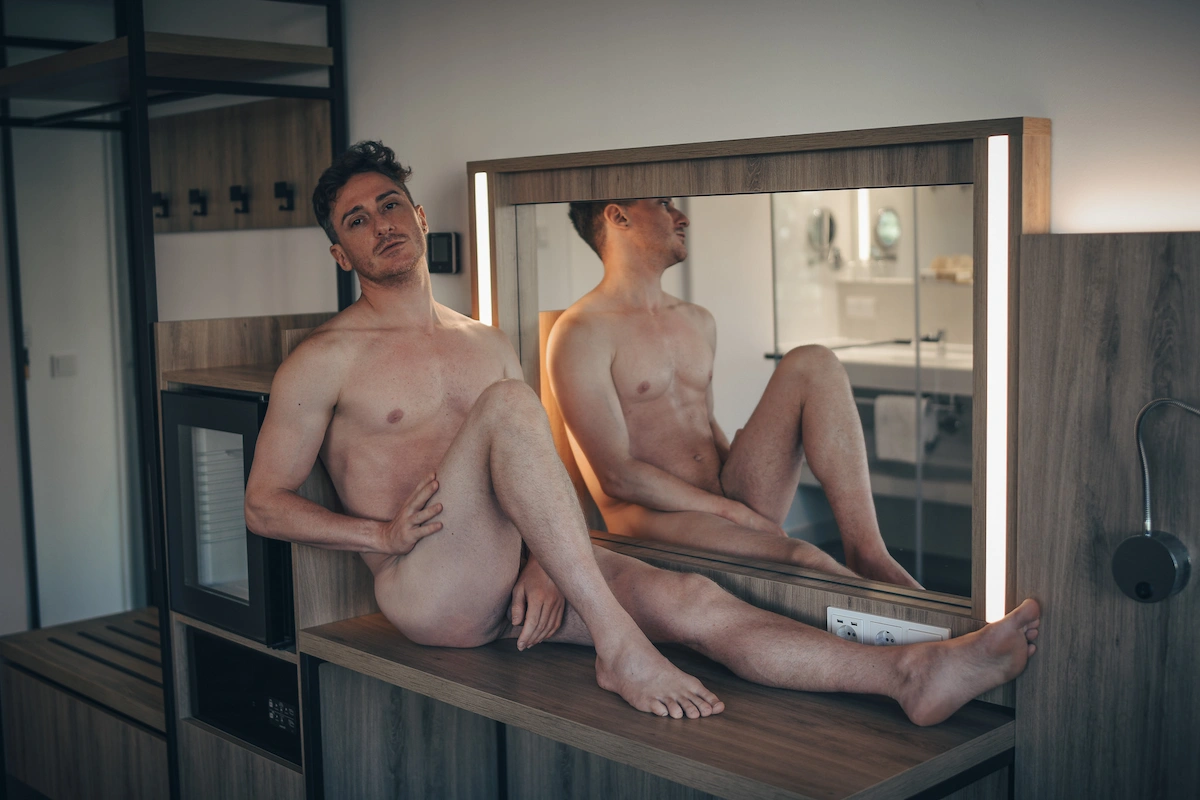 An artistic portrait of a nude man sitting on a wooden counter in front of a backlit mirror in a room, showing his reflection from the profile.