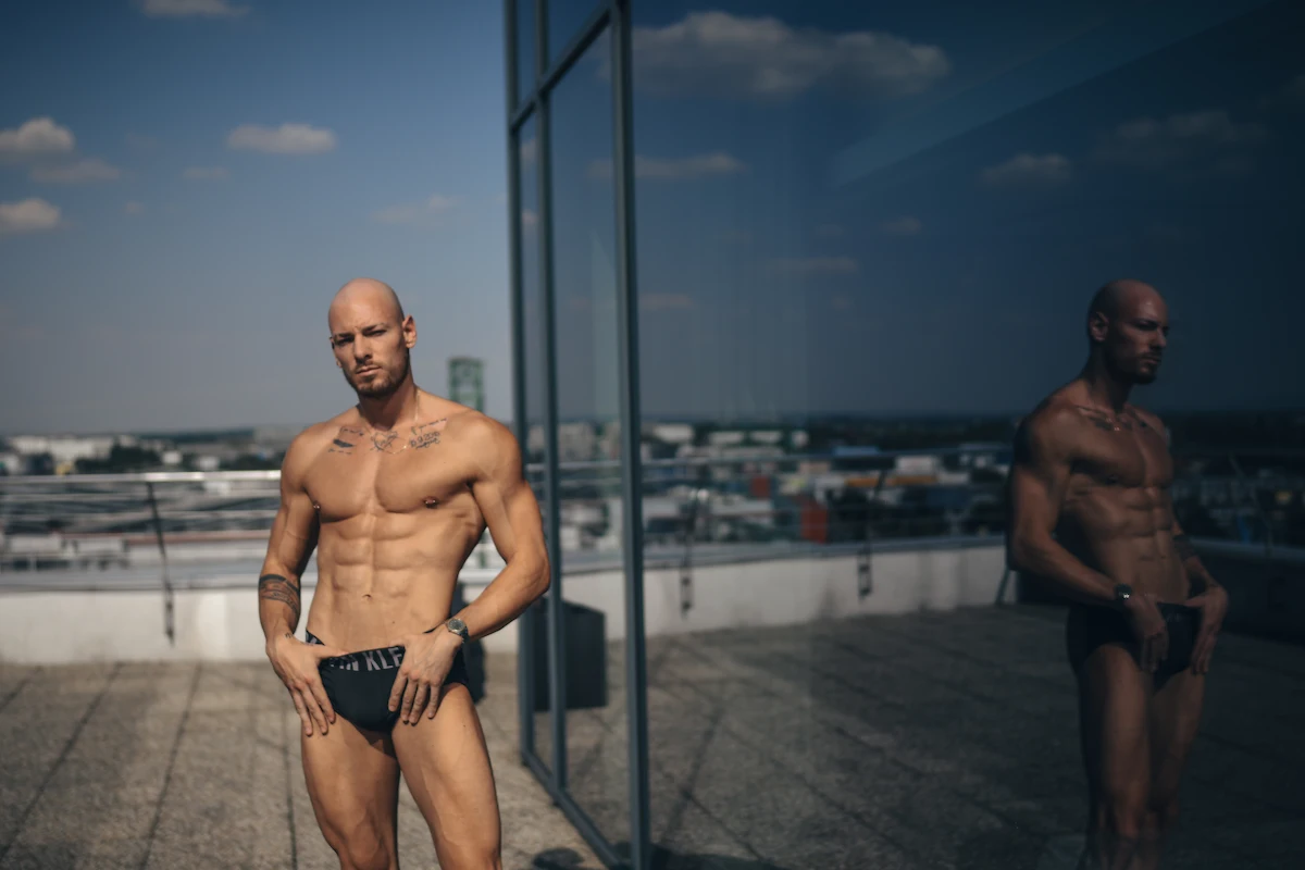 A muscular bald model, Martin S., posing in black underwear on a rooftop terrace in Bratislava. The background features a city skyline under a blue sky, while his detailed silhouette is mirrored in the large glass window next to him.