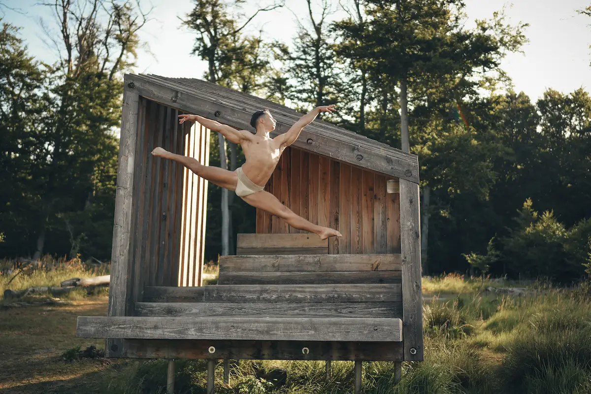 A dynamic shot of a male dancer performing a mid-air split leap inside a modern wooden shelter Vydrica surrounded by trees in daylight close to Bratislava.